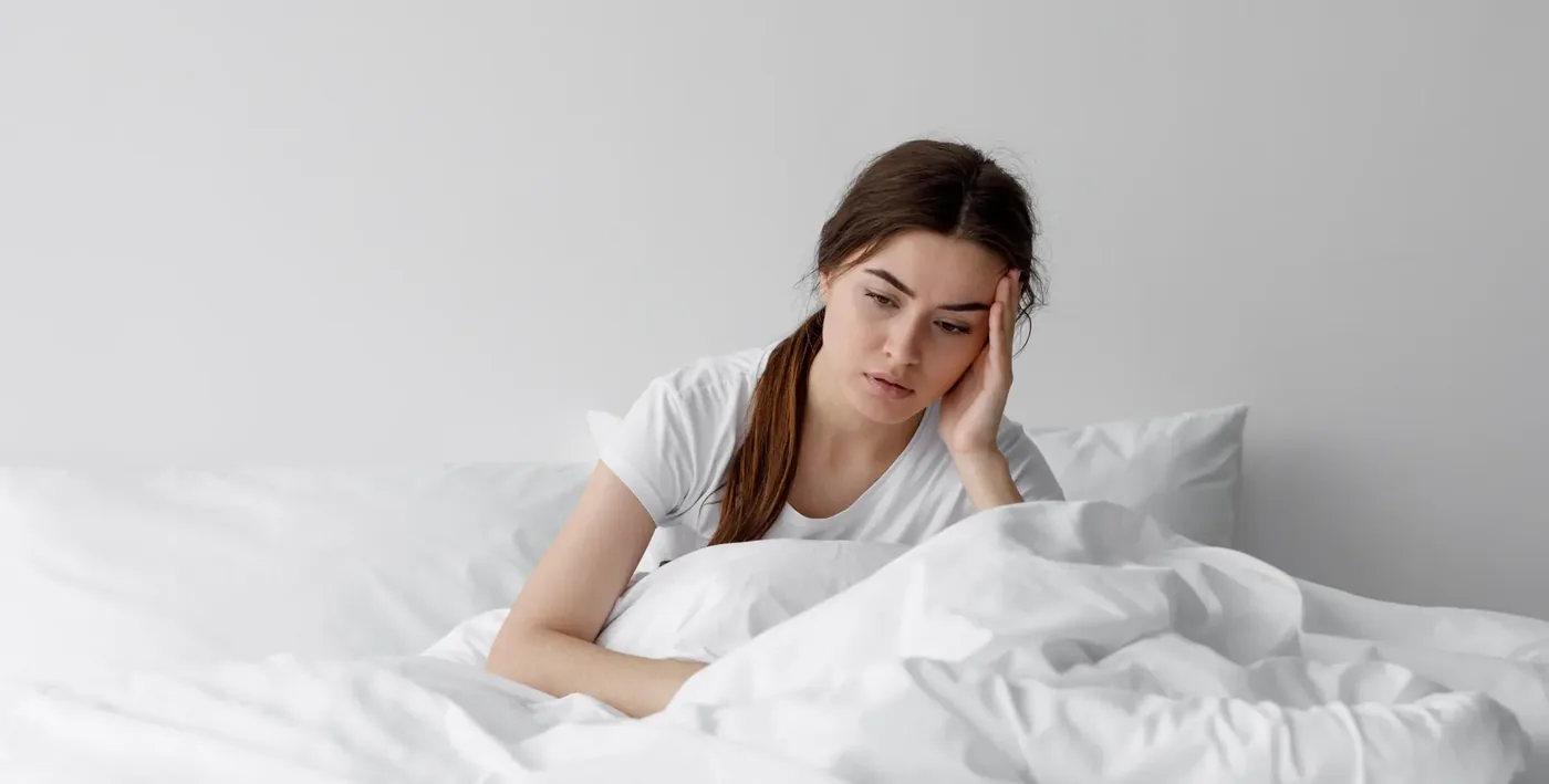 A person sitting up in bed in the morning holding their forehead with a headache
